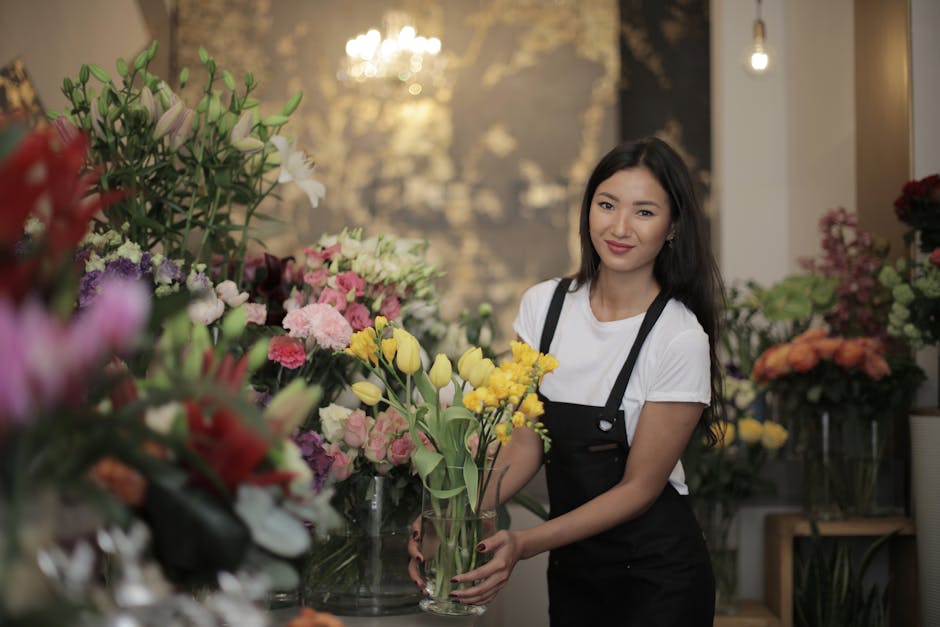 A young female florist with long dark hair and a friendly smile, standing indoors in a flower shop, holding a glass vase of yellow tulips with green stems and leaves. She is dressed in a white t-shirt