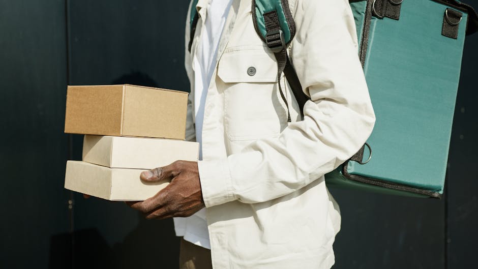 A person wearing a white jacket holding three plain cardboard boxes stacked one on top of the other with their left hand, while their right hand supports the bottom box. The person is also carrying a 