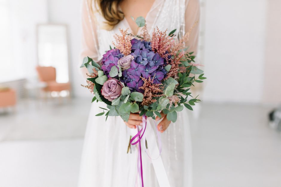 A person dressed in a white wedding gown is holding a large floral bouquet consisting of purple and lavender roses, lavender-hued hydrangeas, and pink astilbe, accented with green eucalyptus leaves. T