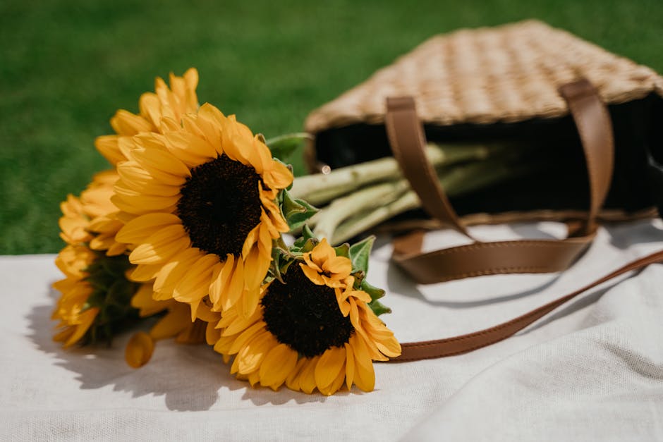 A bouquet of vibrant yellow sunflowers with large, dark brown centers and bright green stems, arranged together and resting on a white cloth surface. The bouquet is placed next to a woven straw bag wi