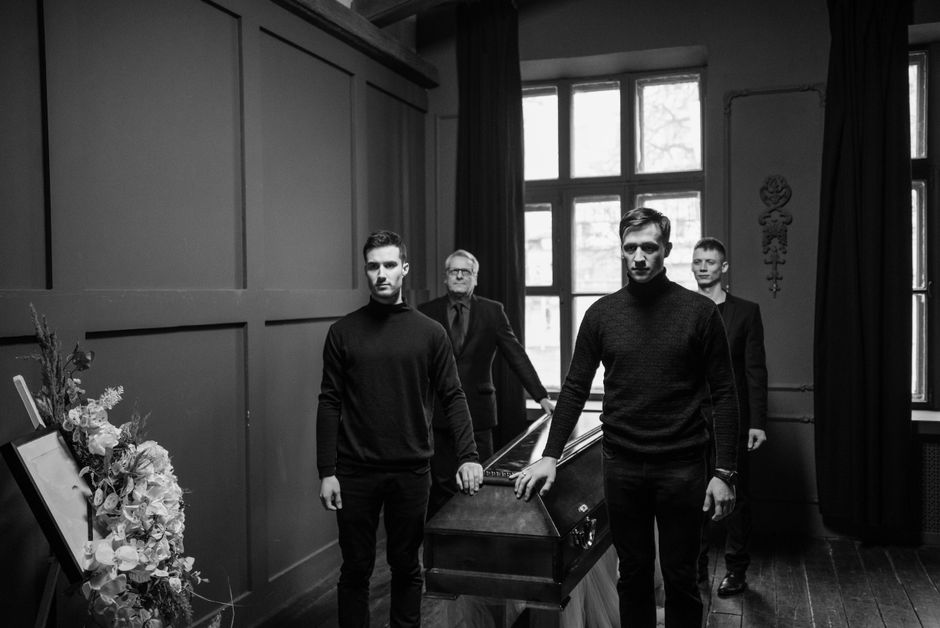 Four men dressed in black, with serious expressions, standing in a dimly lit room with large windows and dark paneling. One man is holding a casket, while the others stand nearby, suggesting a funeral