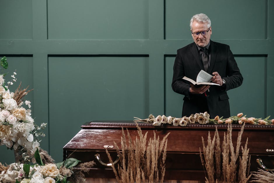 A man dressed in a black suit and glasses stands behind a closed casket adorned with a floral arrangement of white and cream flowers, including anemones, tulips, and roses, with greenery accents. The 