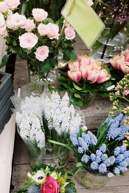 A variety of fresh flower arrangements displayed on a wooden surface, including a bouquet of soft pink roses with green foliage in a clear glass vase, a cluster of white lily of the valley wrapped in 