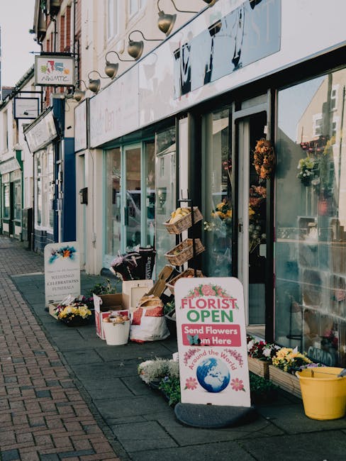 The image depicts the exterior of a flower shop on a busy street, with large glass windows showcasing floral arrangements inside. Outside the shop, there are various flowers and plants displayed on th