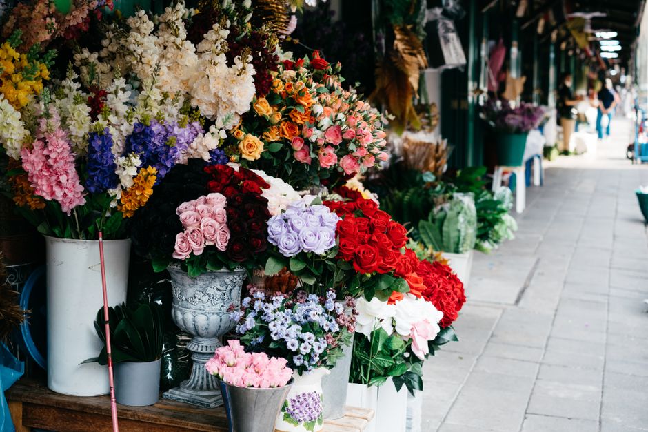A vibrant display of fresh floral arrangements situated outside a flower shop on a paved sidewalk. The flowers include roses, hydrangeas, and various seasonal blooms in colors such as pink, red, purpl